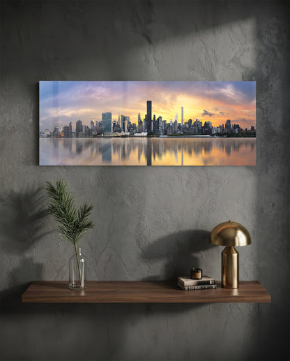 NYC skyline glass wall art mounted above wooden shelf with golden lamp and plant, sunset reflection on water visible.