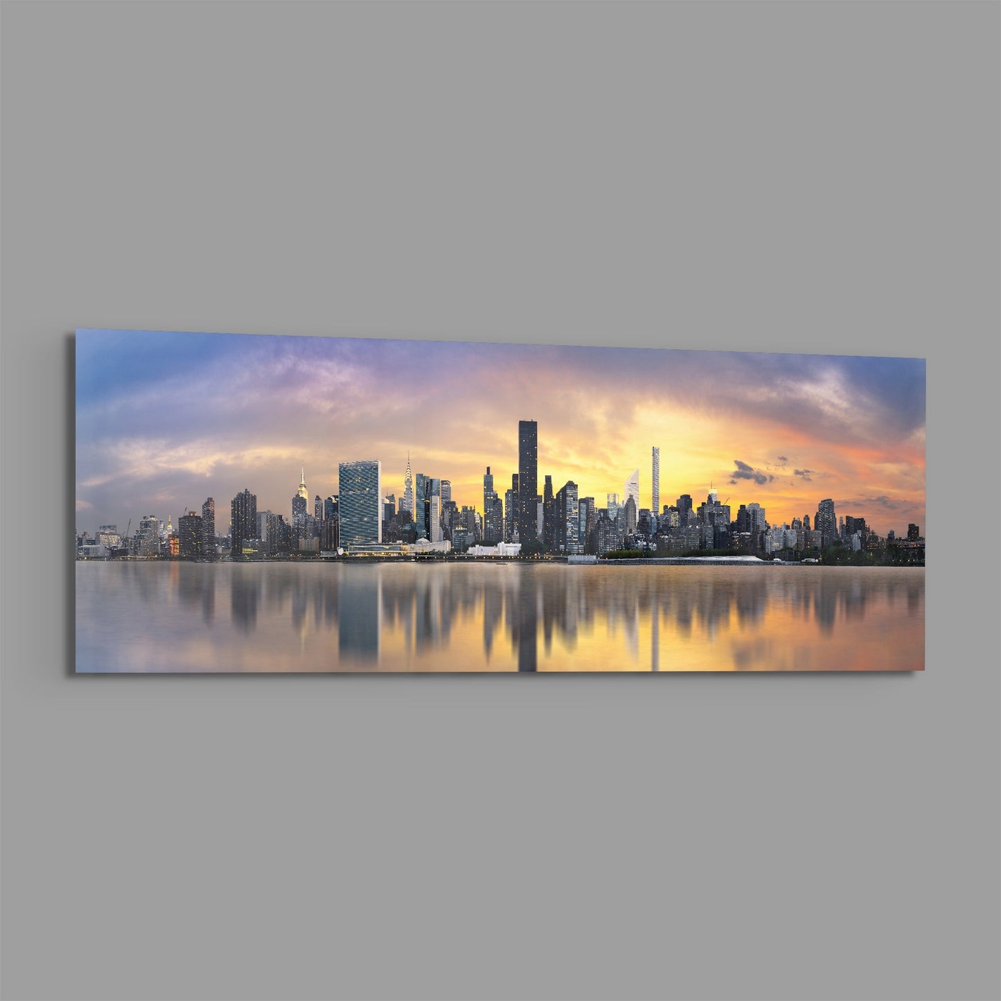 Panoramic glass wall art depicting Manhattan skyline at sunset with golden sky reflected in calm water below.