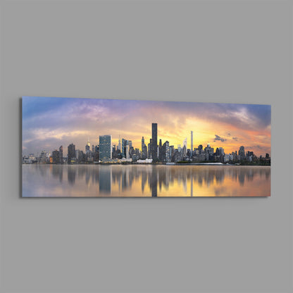 Panoramic glass wall art depicting Manhattan skyline at sunset with golden sky reflected in calm water below.