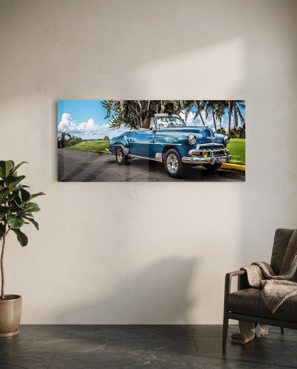 Blue vintage convertible parked under palm trees with green grass and cloudy sky in background.
