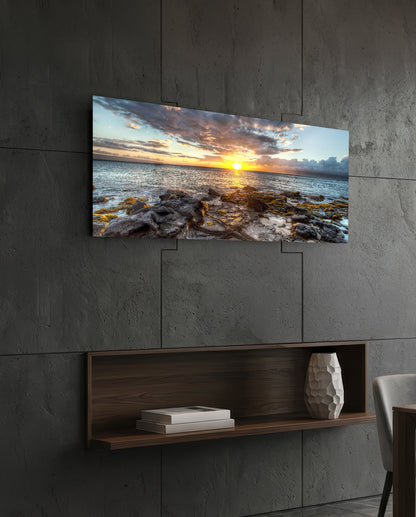 Landscape view of sunset glass wall art mounted above a wooden shelf on a concrete wall, displaying rocky coastline with golden sunset sky.