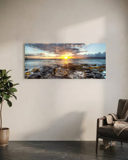 Horizontal glass wall art displaying a coastal sunset with rocky shoreline, golden sun, and dramatic cloudy sky mounted above a leather chair in a modern interior.