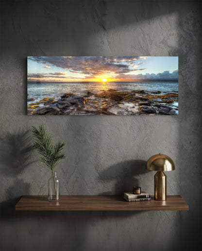 Rectangular glass wall art displaying a sunset over rocky coastline, hung above a wooden shelf with decorative items.