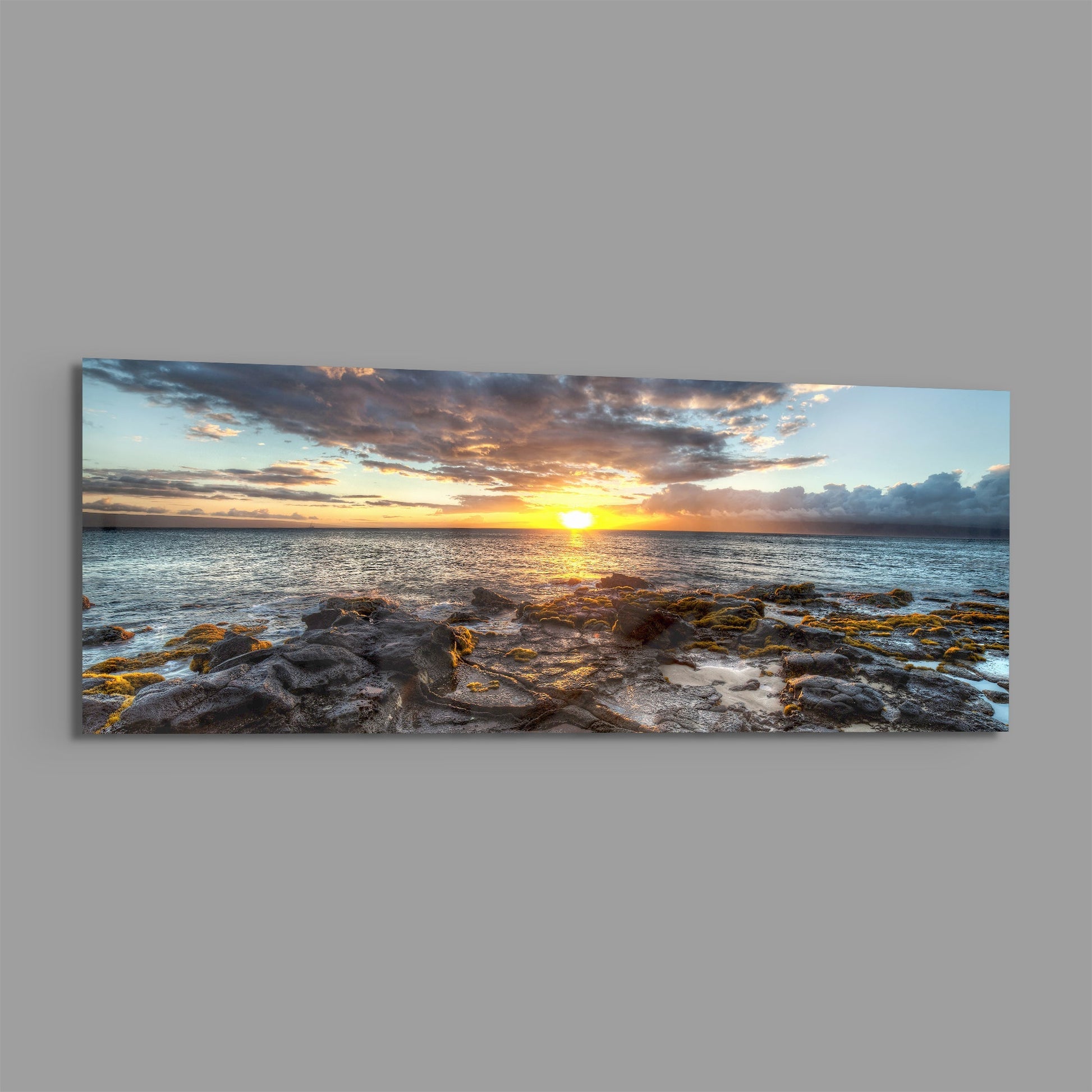 Panoramic glass wall art displaying a golden sunset over ocean waters with rocky shoreline in foreground and dramatic cloudy sky.