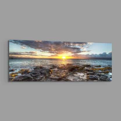 Panoramic glass wall art displaying a golden sunset over ocean waters with rocky shoreline in foreground and dramatic cloudy sky.