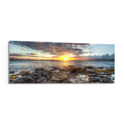 Panoramic glass wall art depicting a golden sunset over rocky coastline with calm ocean waters and dramatic cloudy sky.