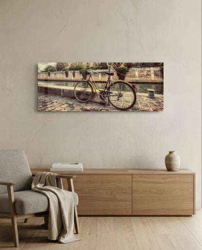 Vintage-style bicycle leaned against stone waterside wall, displayed as horizontal glass wall art above wooden furniture in minimalist room.