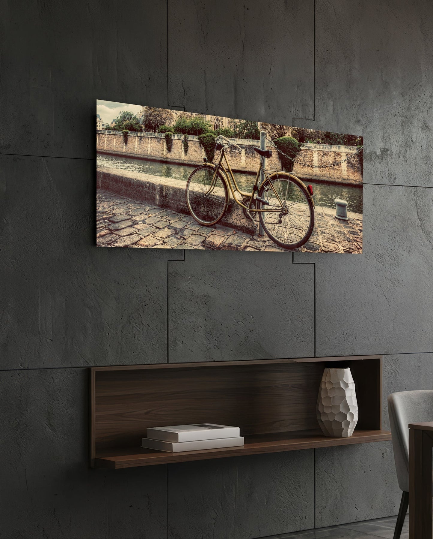 Horizontal glass wall art depicting a vintage bicycle parked against a stone wall beside water, displayed above a wooden shelf.