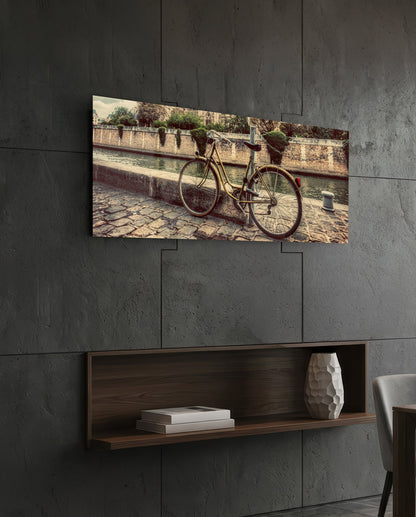 Horizontal glass wall art depicting a vintage bicycle parked against a stone wall beside water, displayed above a wooden shelf.
