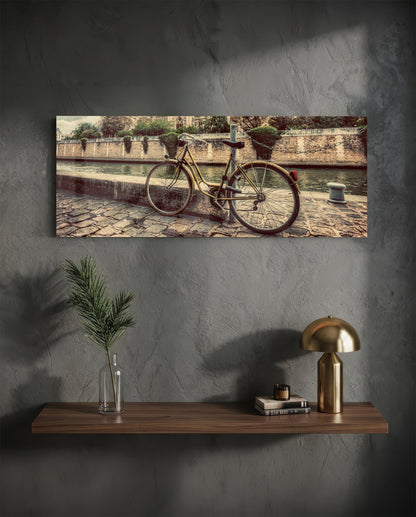 Vintage bicycle leaning against stone waterside wall with sepia-toned architectural background, displayed as horizontal glass wall art above wooden shelf.
