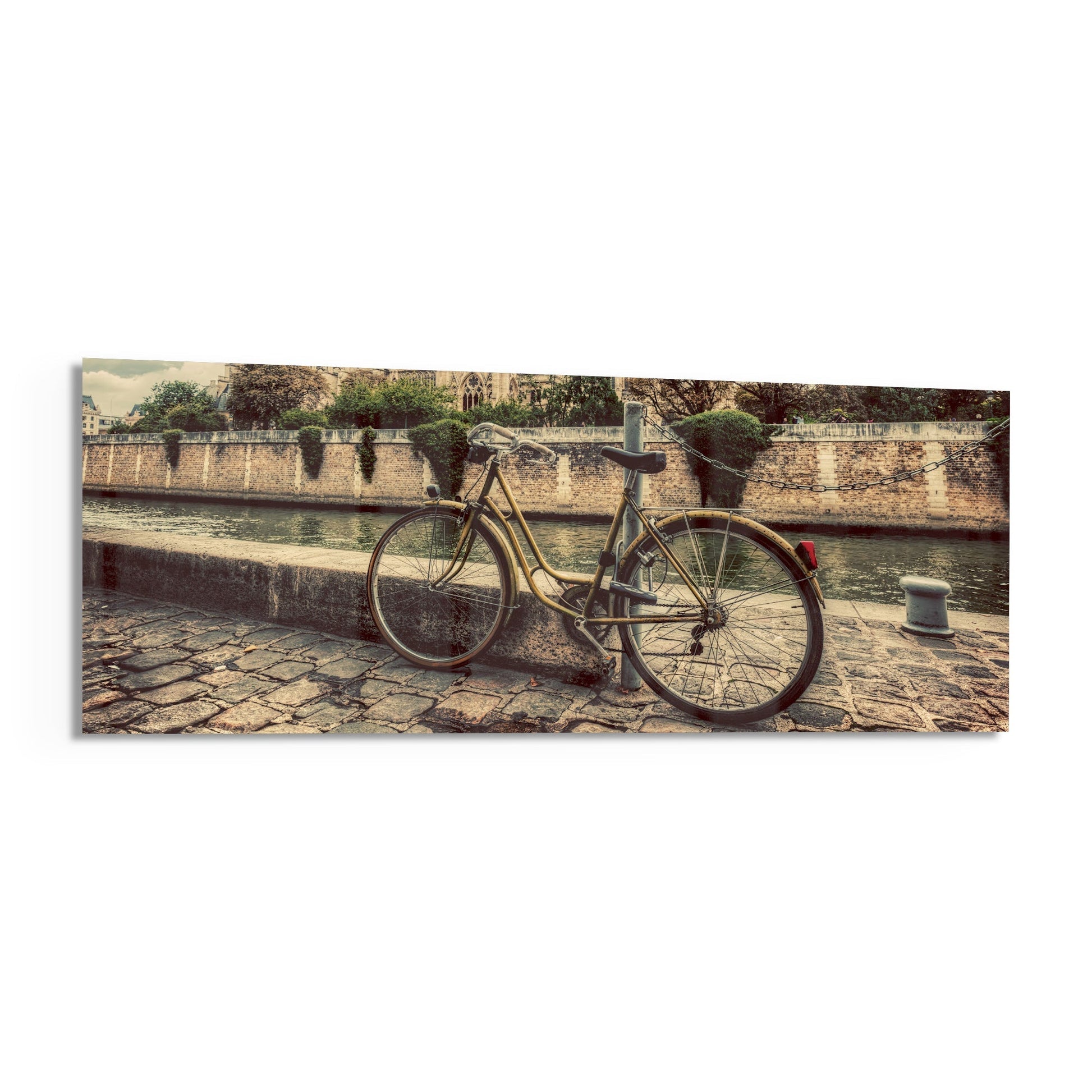 Vintage bicycle with red rear light leaned against stone wall beside calm river, with historic fortification and green trees in background.