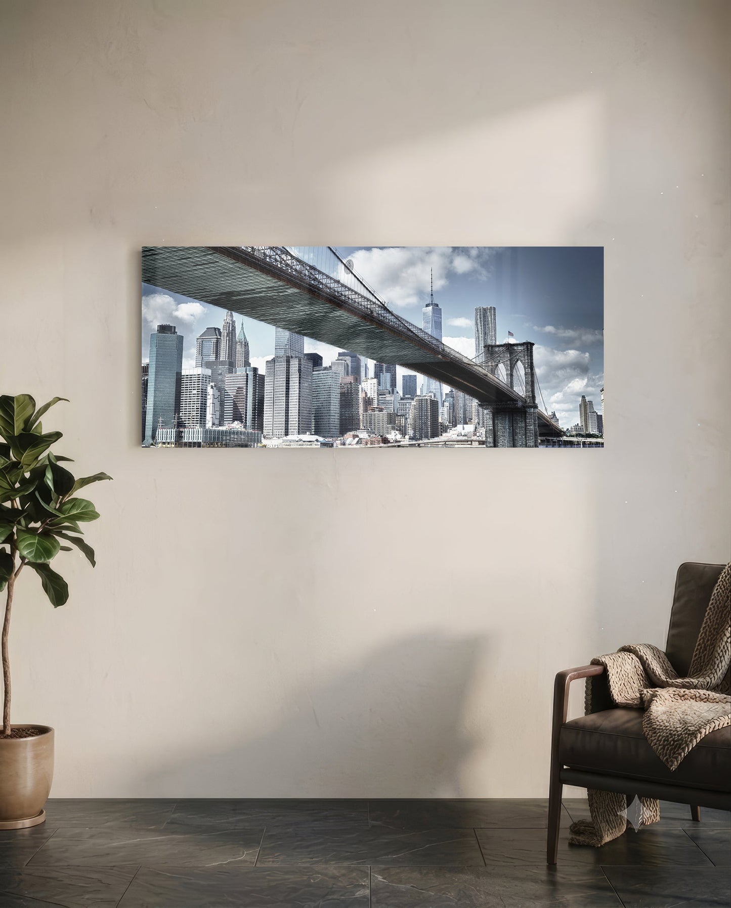 Brooklyn Bridge glass wall art mounted on white wall above leather chair, showing Manhattan skyline and bridge span under blue sky.