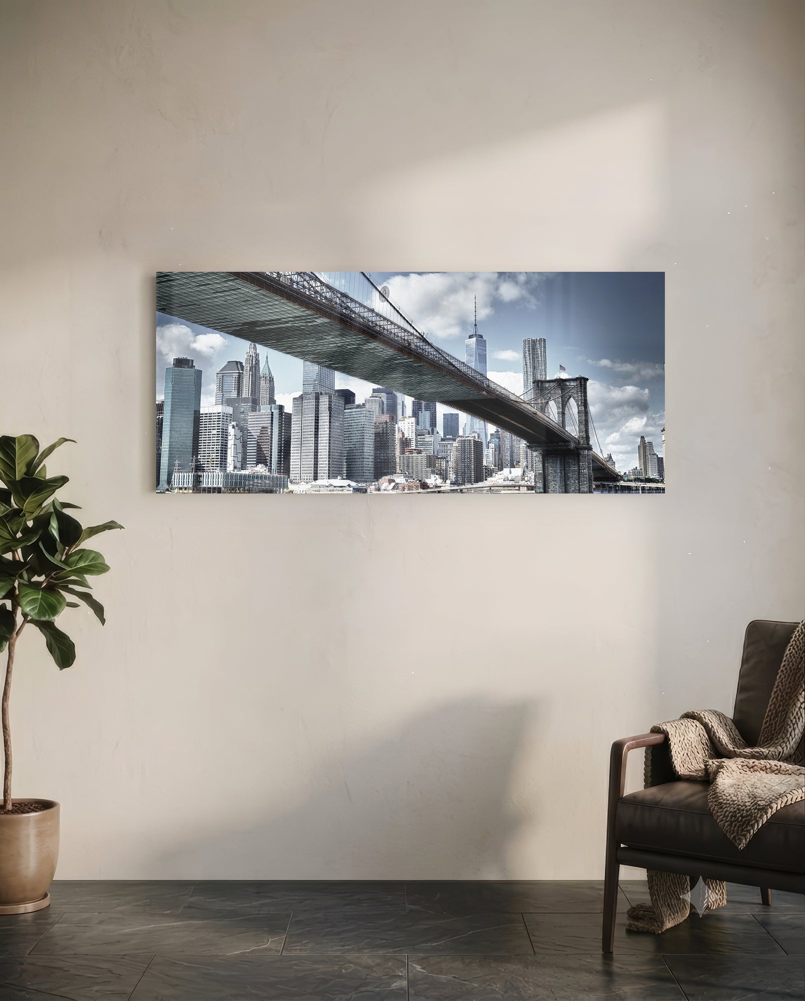 Brooklyn Bridge glass wall art mounted on white wall above leather chair, showing Manhattan skyline and bridge span under blue sky.