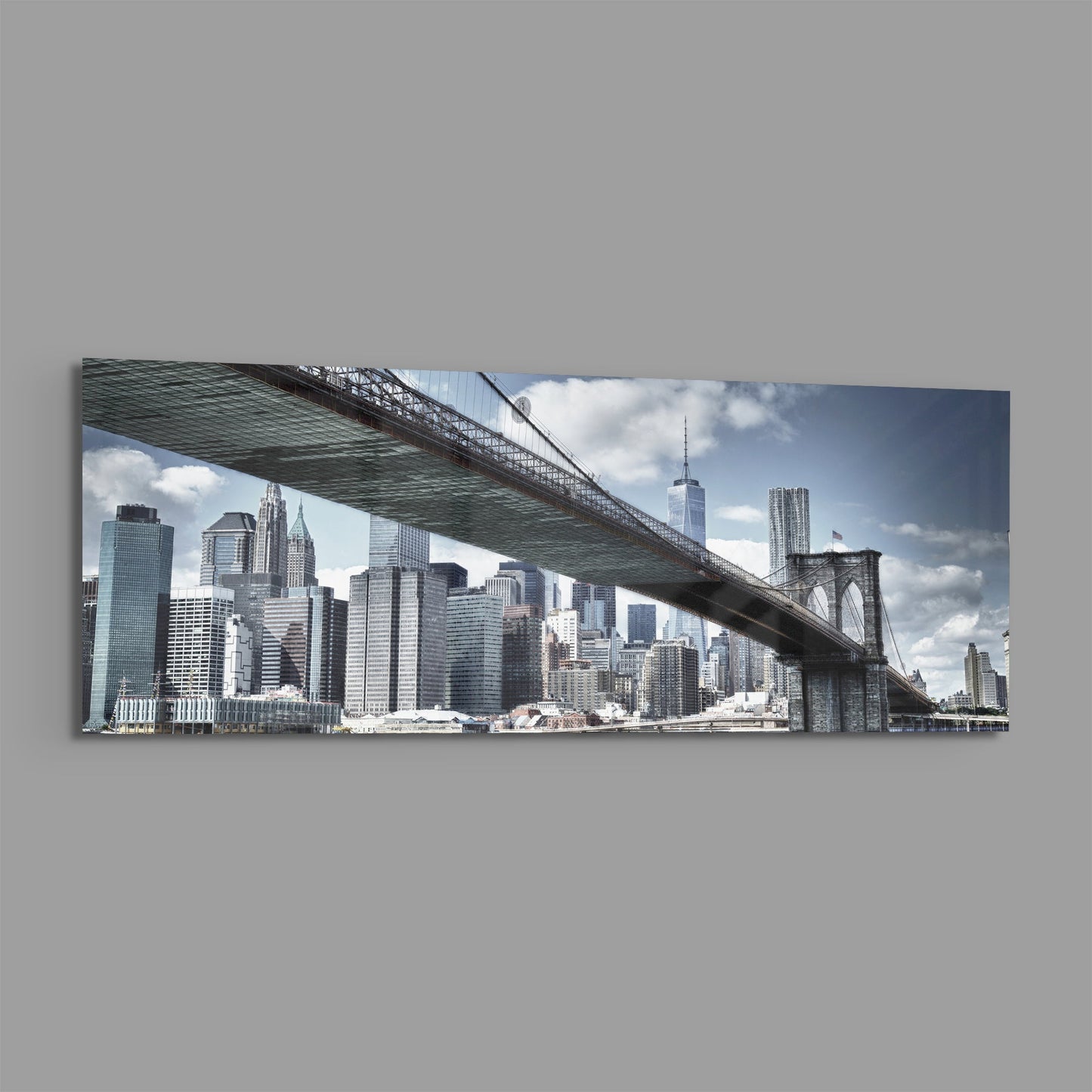 Panoramic glass wall art mounted on gray wall showing Brooklyn Bridge spanning over Manhattan skyline with blue sky and clouds.