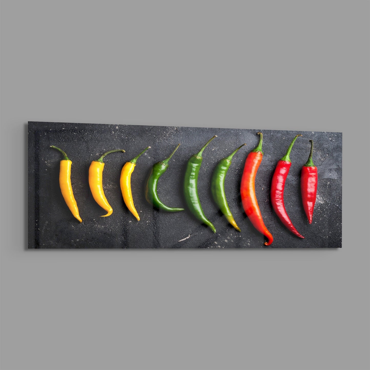 Nine chili peppers in graduating colors from yellow to red arranged in a horizontal line on a dark textured surface.
