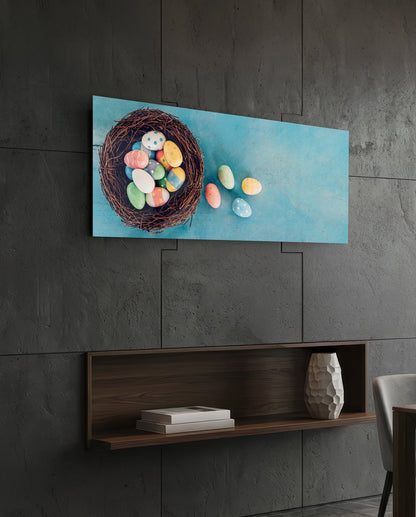 Horizontal glass wall art displaying a woven nest filled with colorful Easter eggs against a light blue background, mounted on a concrete wall above a wooden shelf.