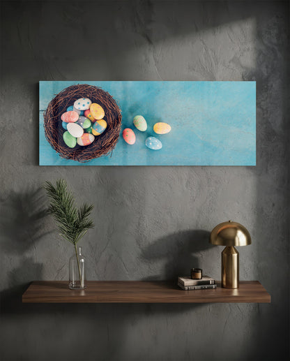 Horizontal glass wall art displaying colorful painted Easter eggs spilling from a woven nest against a blue background, installed above a wooden shelf.