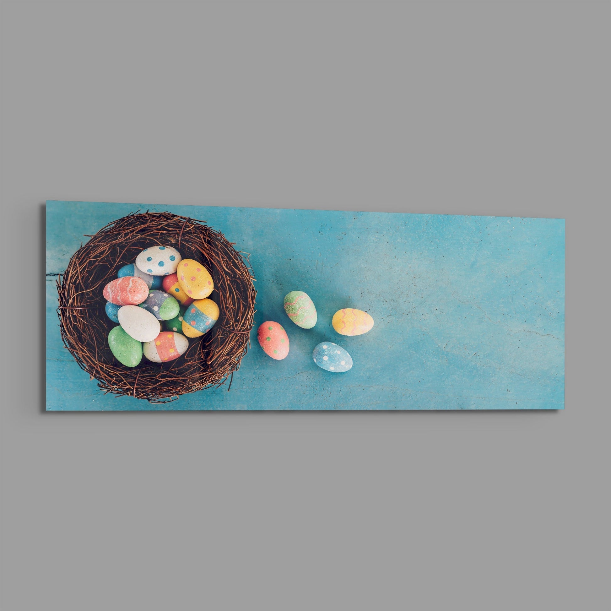 Hand-painted decorative Easter eggs spilling from a brown twig nest onto a textured turquoise background on rectangular glass wall art.