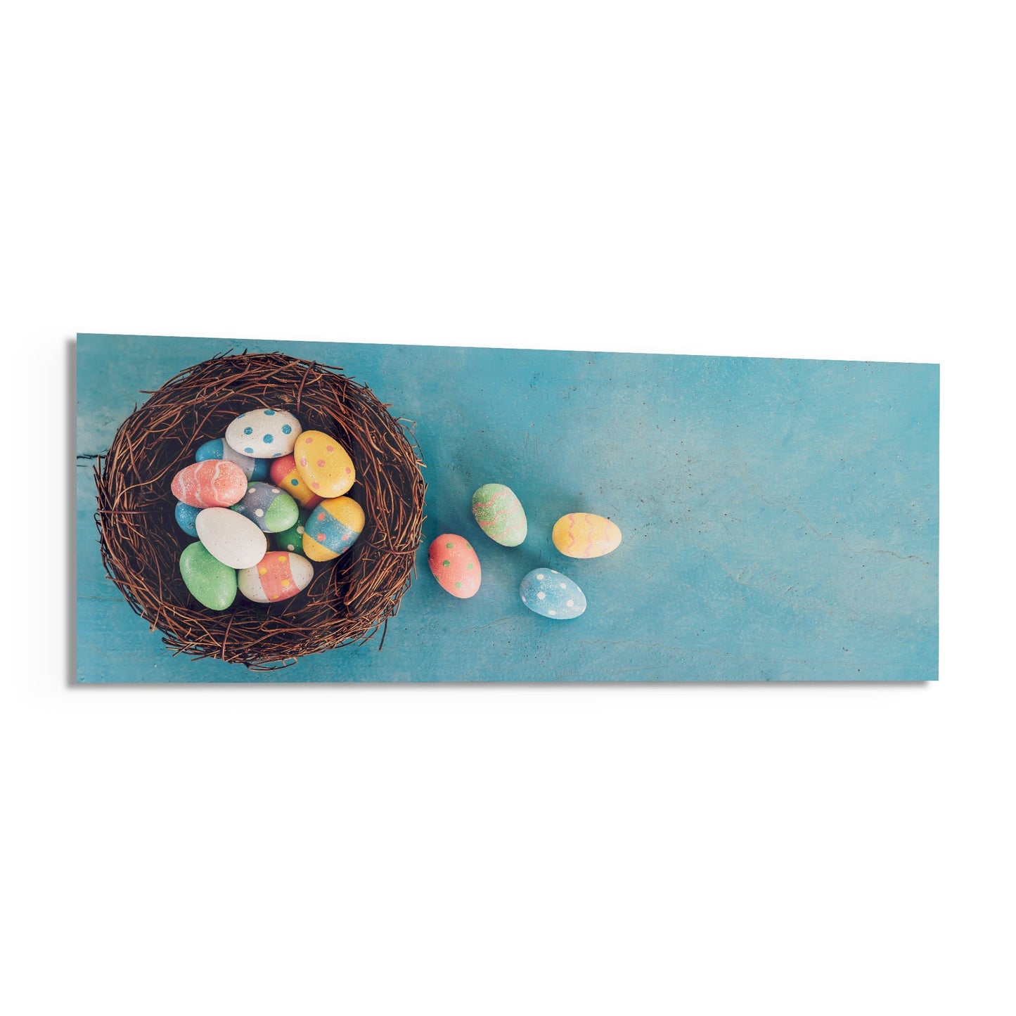 Decorated Easter eggs spilling from a brown woven nest against a turquoise blue background on rectangular glass wall art.