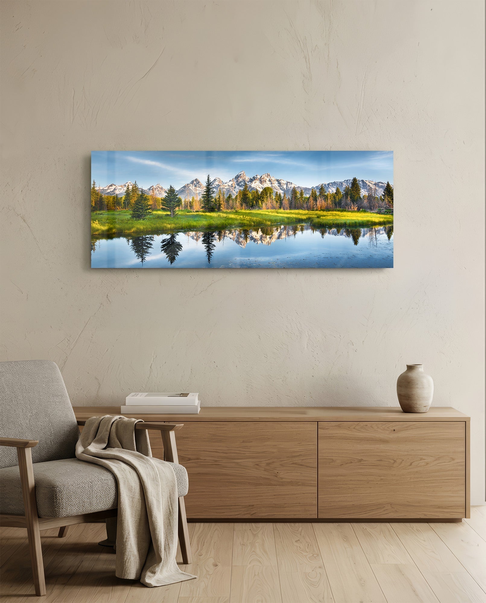 Horizontal glass wall art displaying a mountain landscape with snow-capped peaks, evergreen trees, and their reflections in a still river, mounted above a wooden console table.