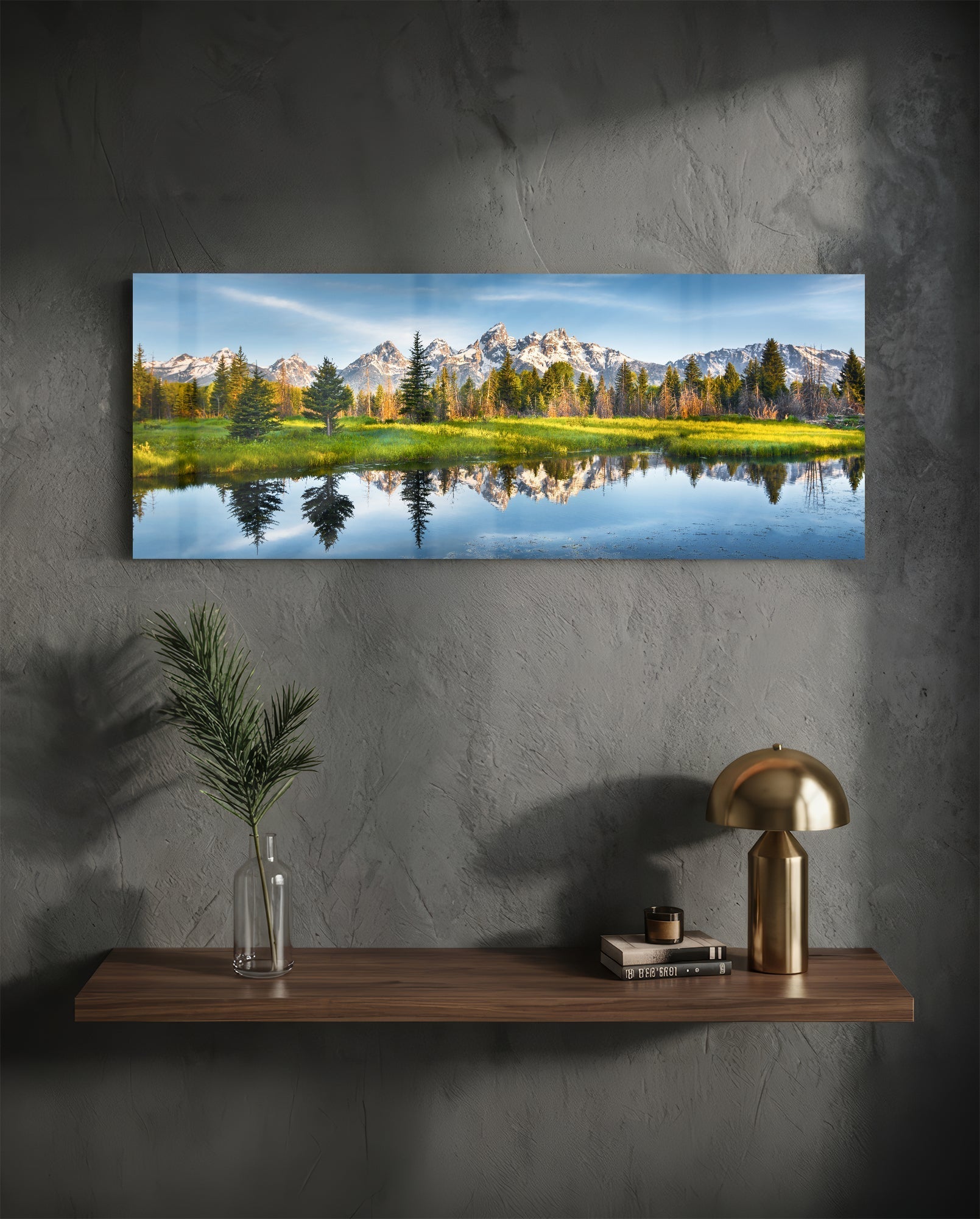 Horizontal glass landscape print showing mountains, forest, and water reflection mounted on gray concrete wall above wooden shelf with brass lamp.