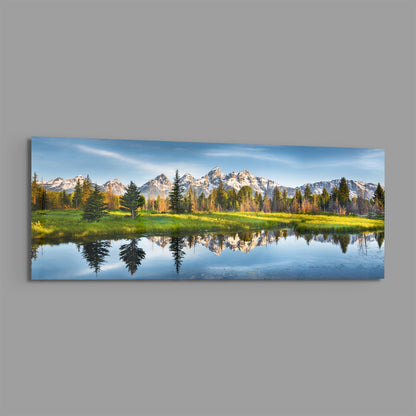 Snow-capped mountain range reflected in still water surrounded by green meadow and evergreen forest under blue sky on glass wall art.