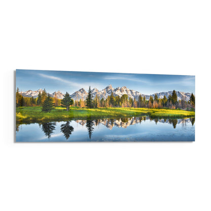 Snow-capped mountain range reflected in still water with evergreen forest and green meadow under blue sky on glass wall art.
