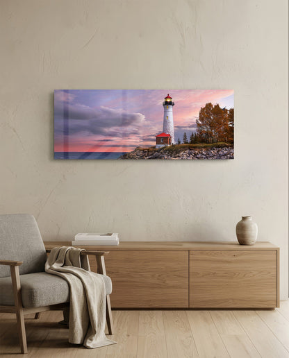 Horizontal glass wall art displaying a white lighthouse with red base at sunset, mounted above wooden furniture in a minimalist room.