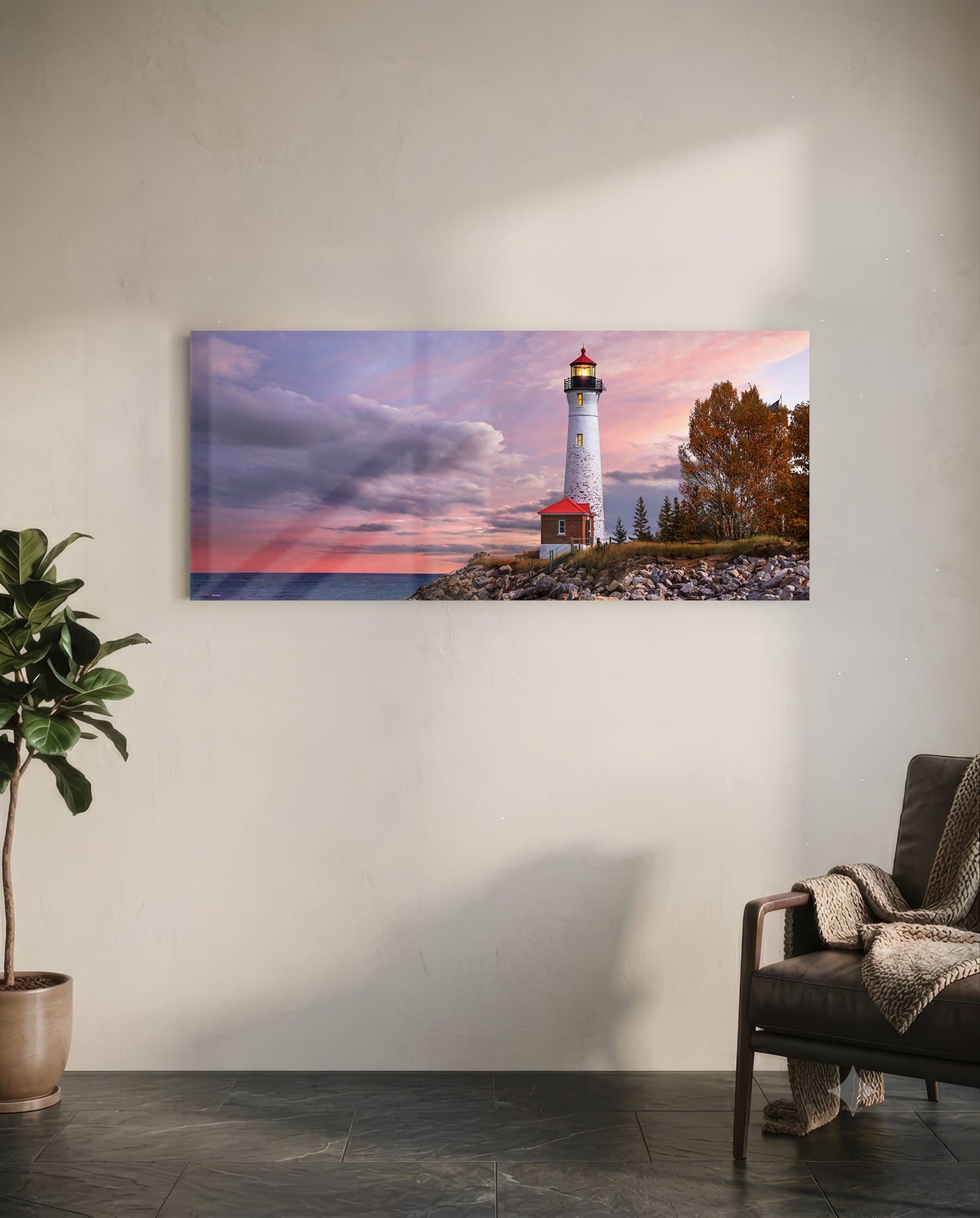 Landscape glass wall art displaying a white lighthouse with red roof at sunset, hung on white wall above leather chair and potted plant.
