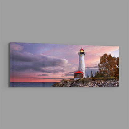 White lighthouse with red base on rocky shoreline at sunset, purple and pink sky, autumn trees, calm water, panoramic view.
