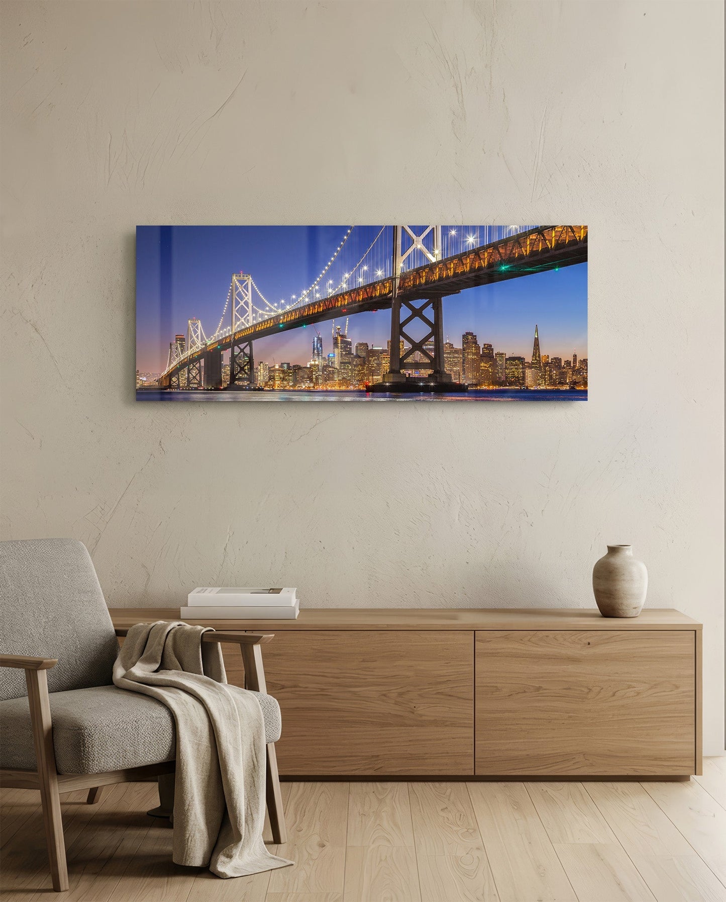 Illuminated Bay Bridge and San Francisco skyline at twilight displayed as horizontal glass wall art above wooden dresser in modern living room.