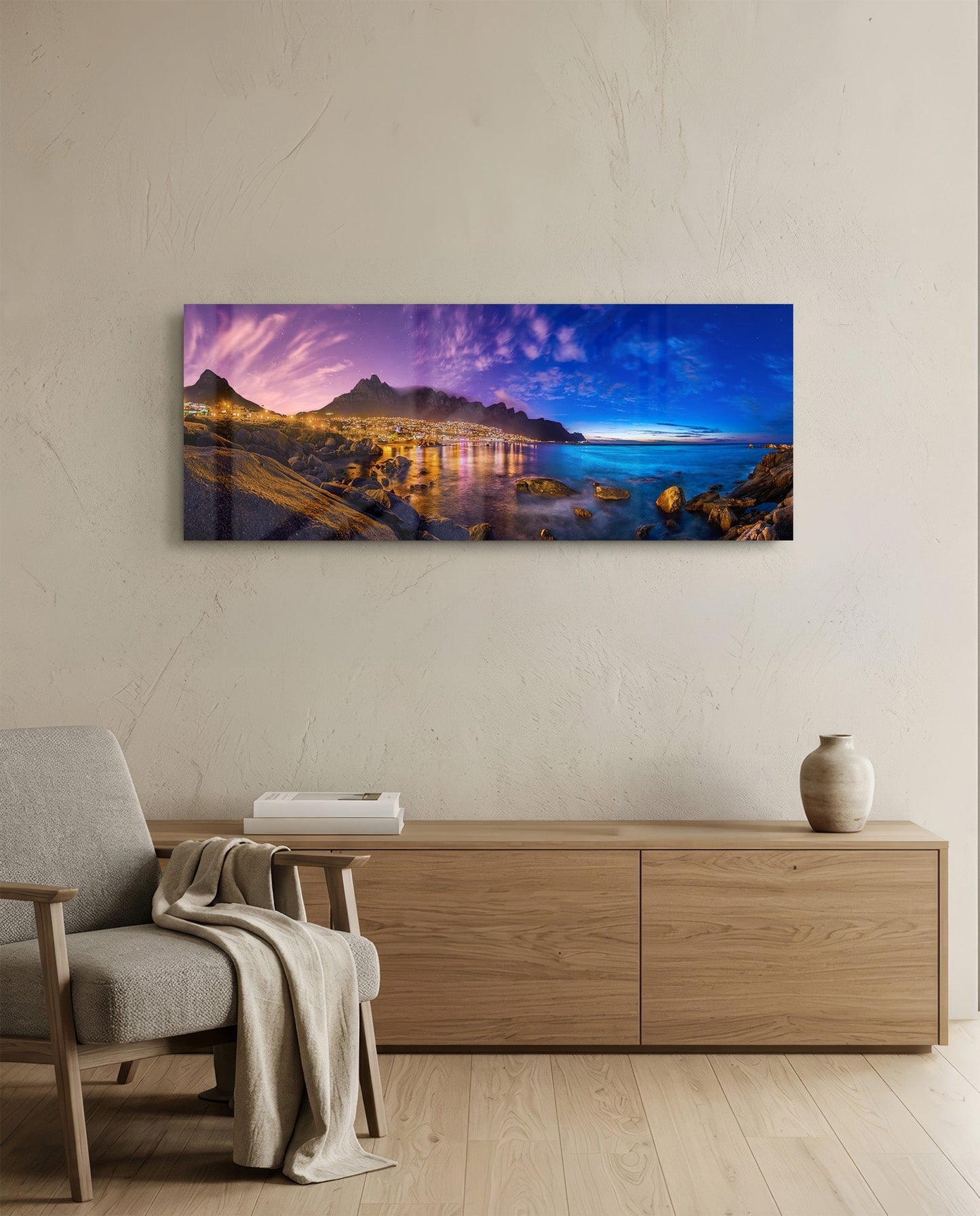 Glass wall art displaying a twilight coastal landscape with illuminated city, mountains, and blue ocean mounted above a wooden console table in a minimalist interior.