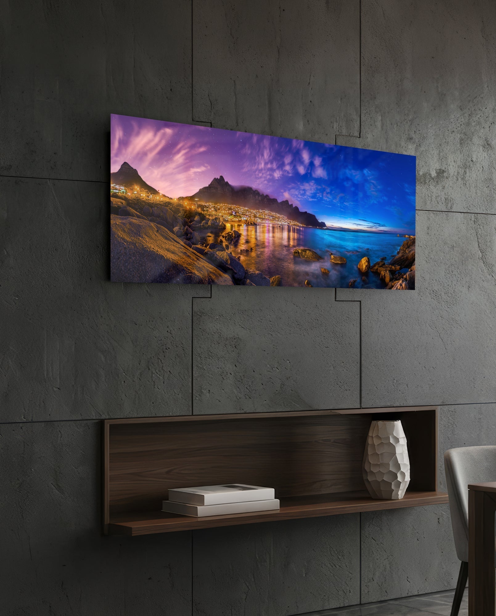 Horizontal glass wall art depicting a twilight coastal landscape with illuminated mountains, water, and rocks mounted on concrete wall above wooden shelf.