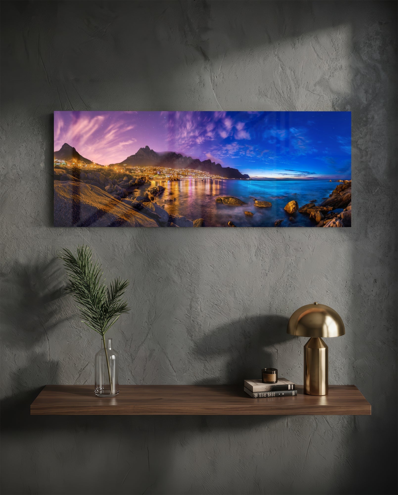 Landscape glass wall art depicting twilight coastal scene with illuminated mountainside town, blue ocean, and rocky foreground, displayed above wooden shelf.