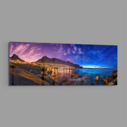 Panoramic glass wall art displaying a twilight coastal scene with illuminated mountainside town, calm blue water, and rocky foreground at dusk.
