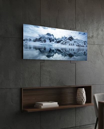 Rectangular glass wall art mounted on concrete wall above wooden shelf, displaying snow-capped mountains reflected in still water under cloudy sky.