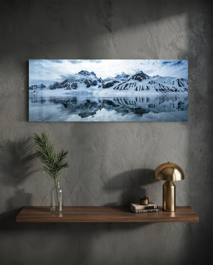 Glass wall art hanging above wooden shelf displaying snow-capped mountains reflected in still water.