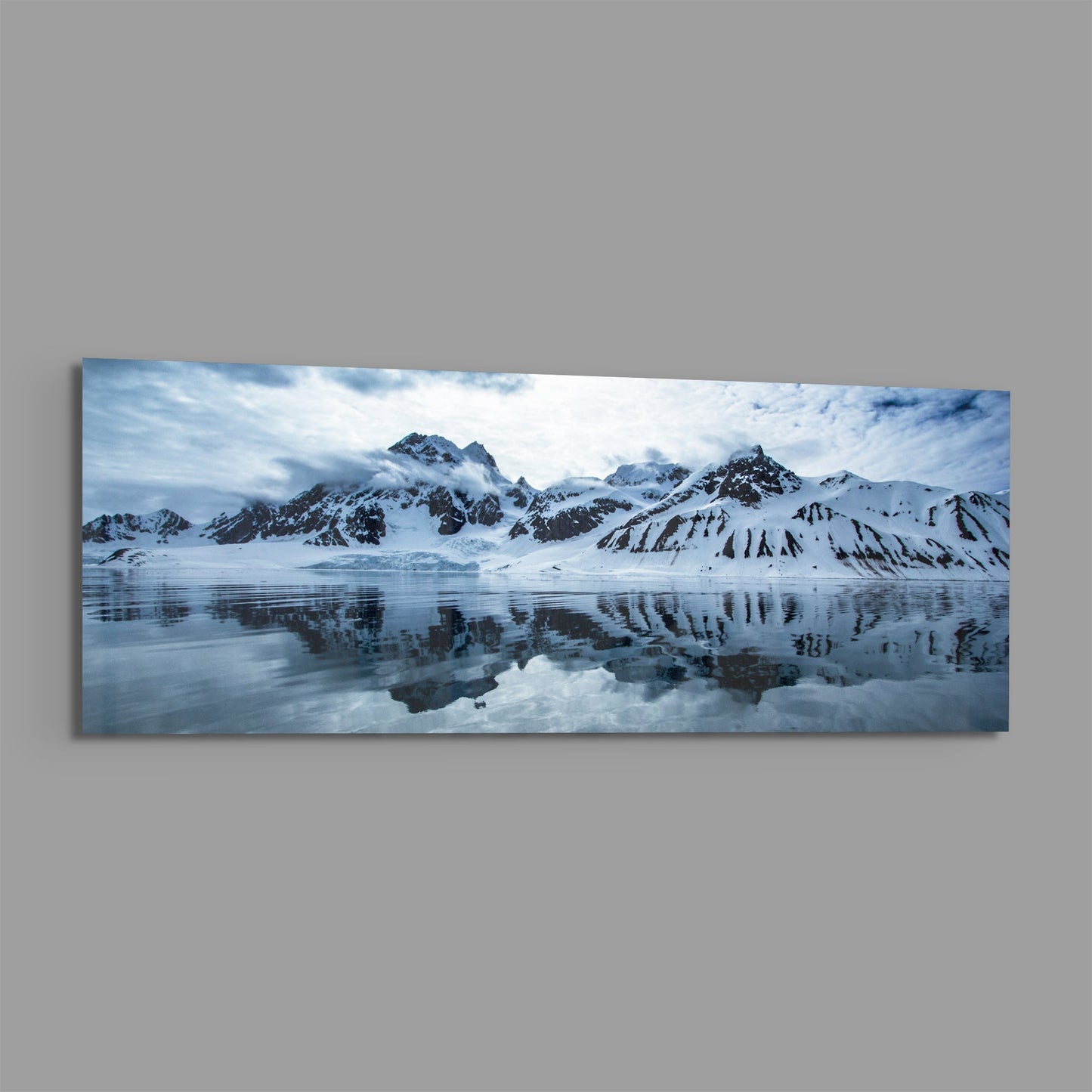 Snow-covered mountains reflected in still icy water under cloudy sky, panoramic landscape photograph on glass.