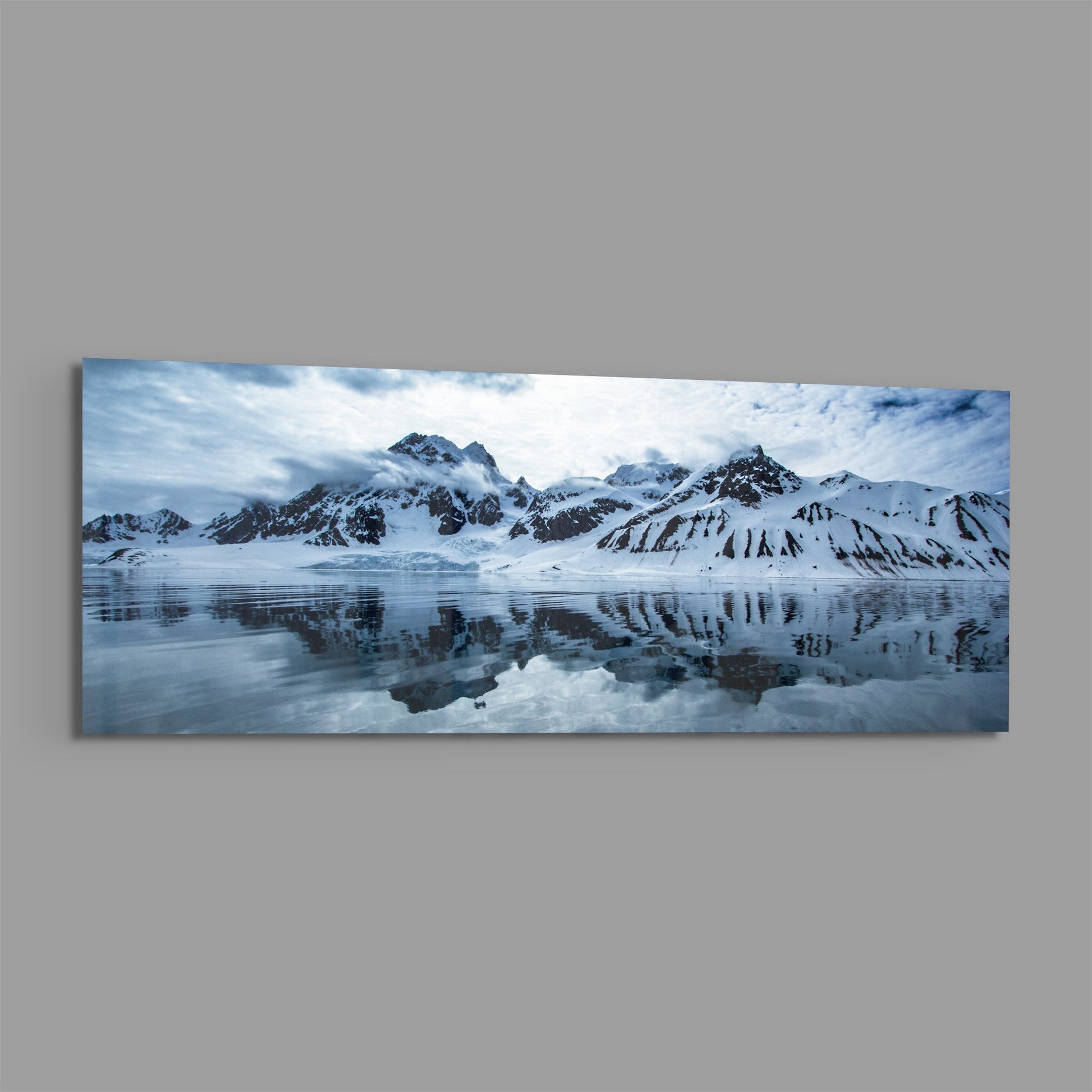 Snow-covered mountains reflected in still icy water under cloudy sky, panoramic landscape photograph on glass.