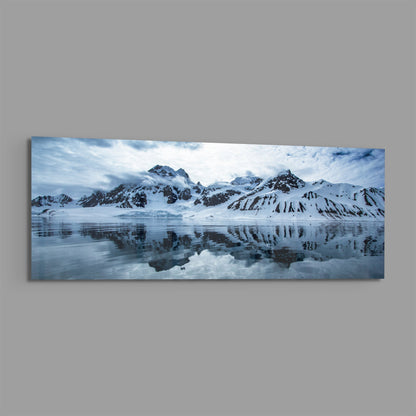 Snow-covered mountains reflected in still icy water under cloudy sky, panoramic landscape photograph on glass.