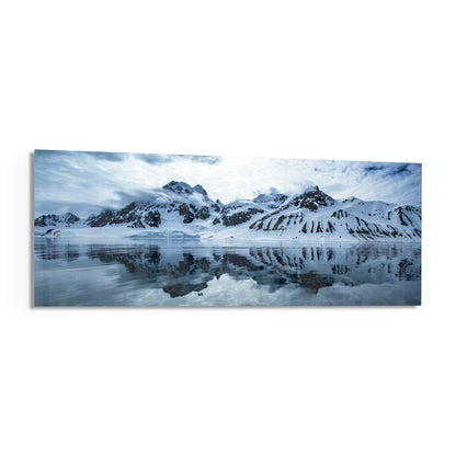 Panoramic glass wall art depicting snow-covered mountains reflected in still icy water under cloudy sky.