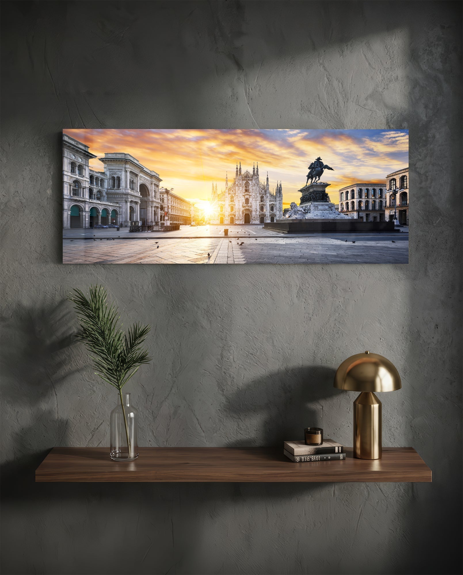 Horizontal glass wall art displaying Milan's Duomo cathedral at golden sunset, mounted above wooden shelf with brass lamp and plant vase.