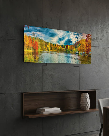 Horizontal glass wall art displaying autumn lakeside landscape with colorful fall trees, blue water, and sky, mounted on concrete wall above wooden shelf.