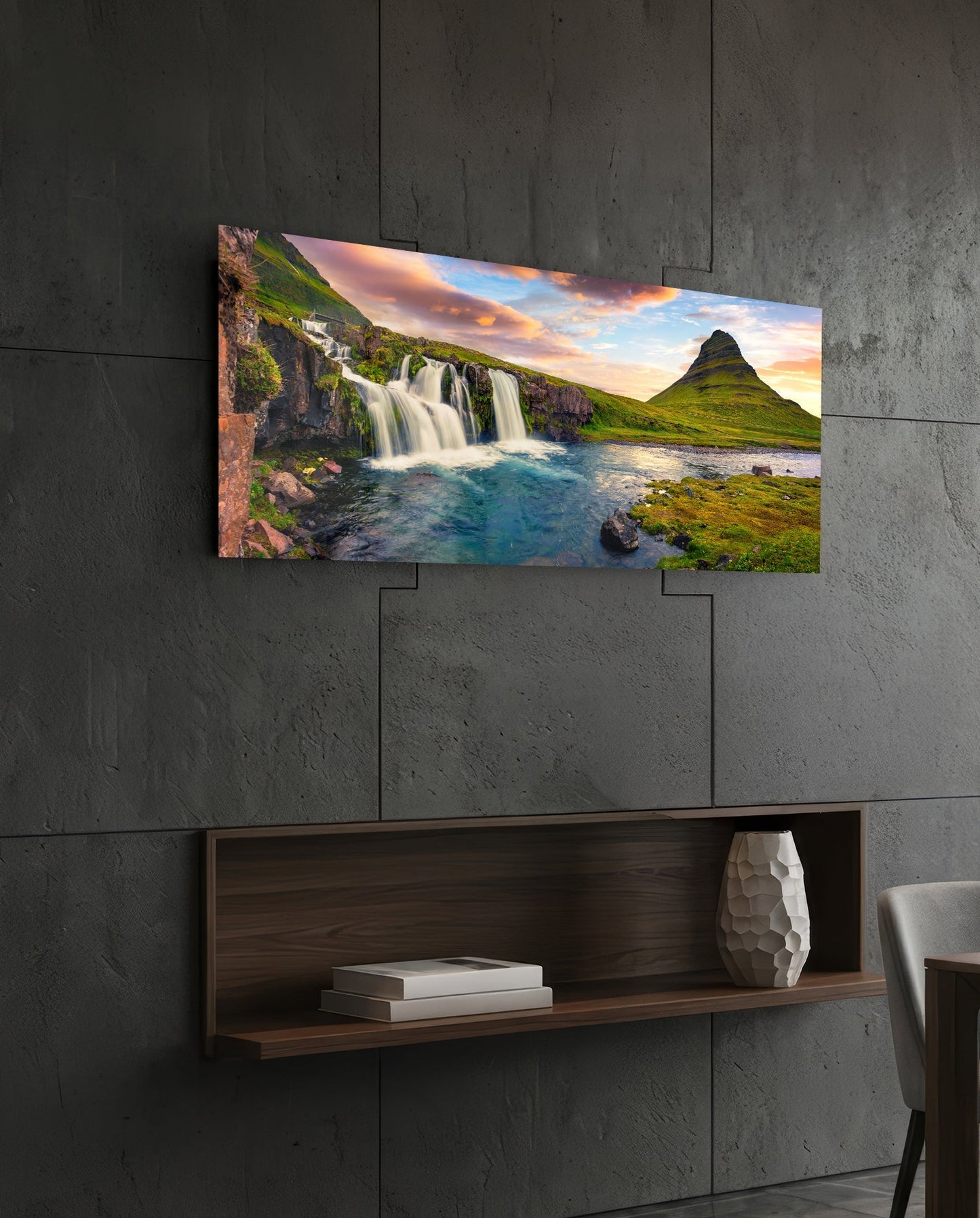 Rectangular glass wall art displaying an Icelandic waterfall landscape with green mountains and sunset sky, mounted on a concrete wall above a wooden shelf.
