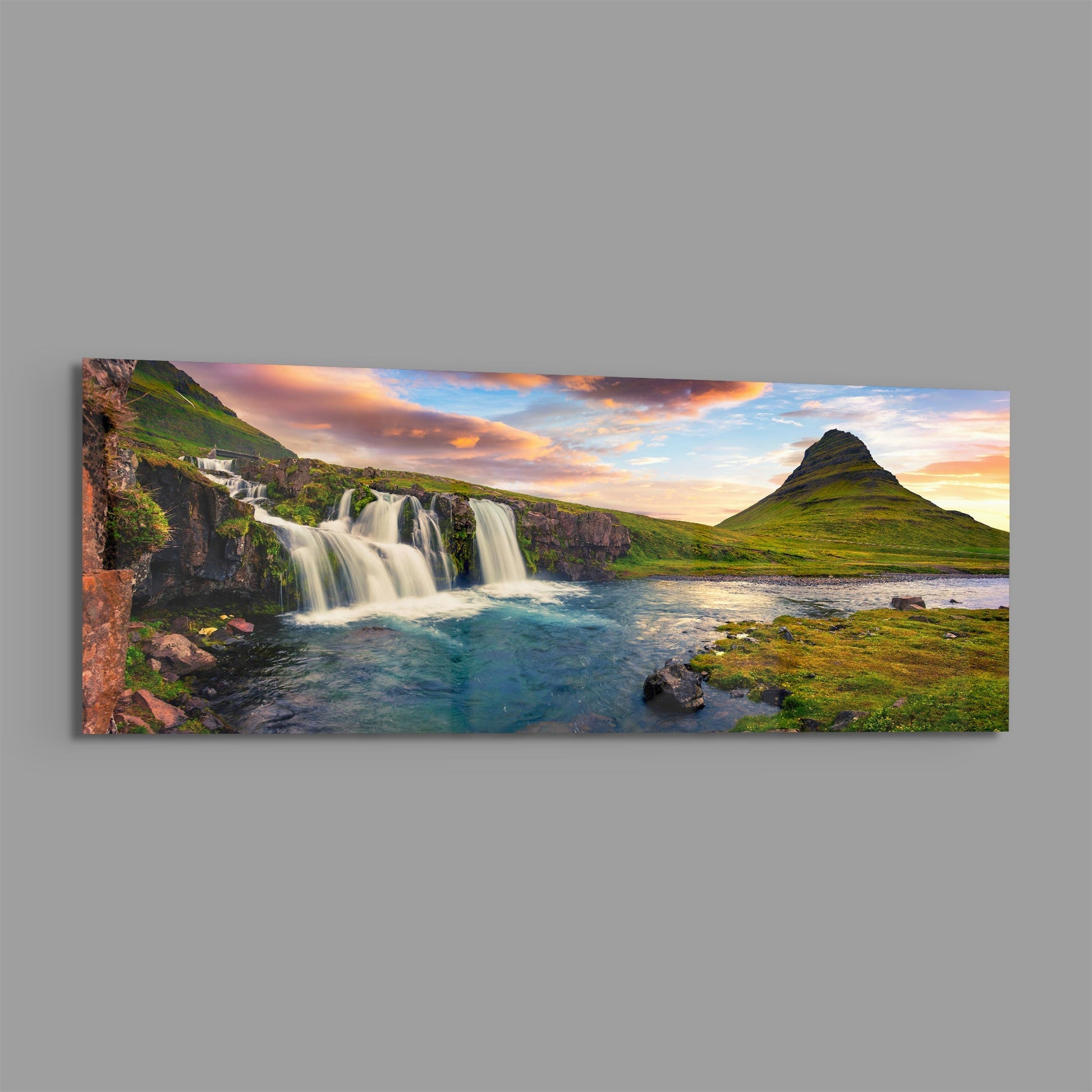 Panoramic glass wall art displaying Icelandic waterfall cascading into turquoise river with green mountain peak at sunset.