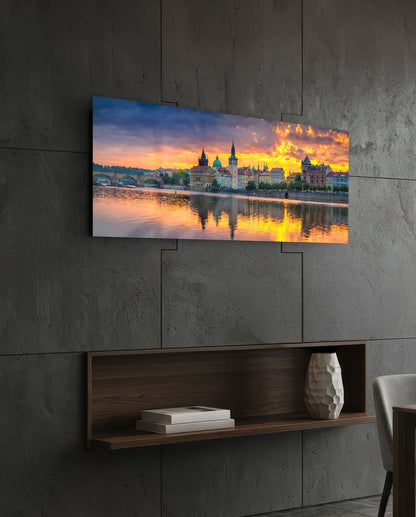 Panoramic glass wall art displaying a sunset over Prague's riverside architecture, mounted on concrete wall above wooden shelf.