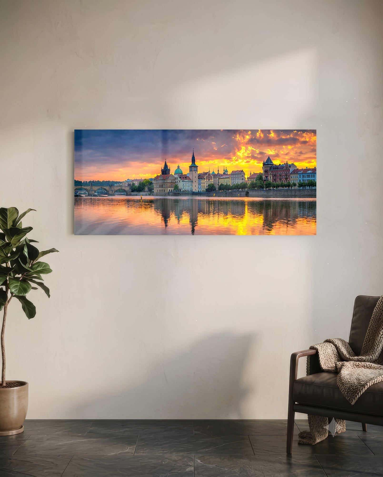 Prague Glass Wall Art displayed on white wall above leather chair, showing sunset cityscape with golden reflections on water.