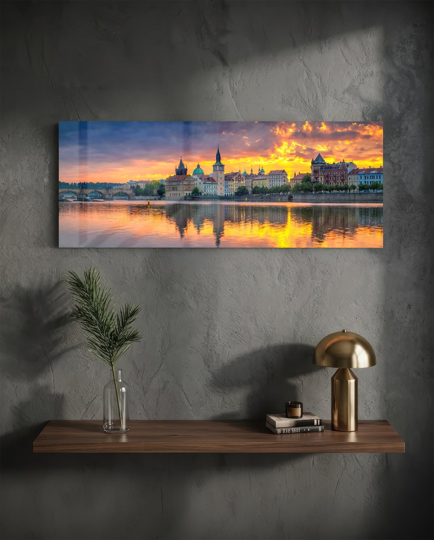 Horizontal glass wall art displaying Prague's riverside architecture at sunset, hung above wooden shelf with brass lamp and decorative items.