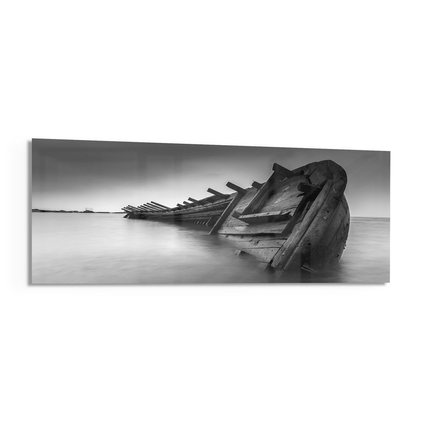 Monochrome photograph of a deteriorated wooden shipwreck partially submerged in calm shallow water, displaying exposed timber ribs and skeletal structure extending into the distance.