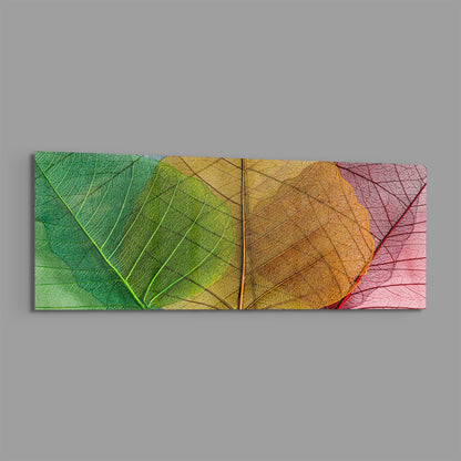 Horizontal glass wall art panel displaying three overlapping translucent leaves in green, yellow-gold, and pink tones against gray background.
